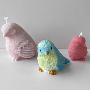 May include: Three bird-shaped candles in different sizes and colours. A large pink candle, a medium blue and green candle, and a small red candle. The candles have detailed feather textures and are on a white surface.