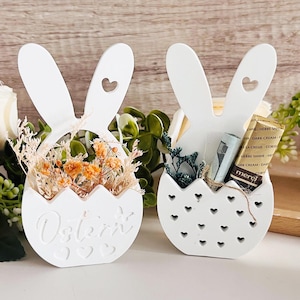 May include: Two white wooden bunny-shaped Easter baskets. One basket is filled with dried flowers and the other with a small gift wrapped in brown paper and a chocolate bar. Both baskets have a heart-shaped cutout on the ears and a scalloped edge around the bottom.