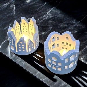 May include: Two white ceramic candle holders with intricate cut-out designs resembling buildings. The candle holders are lit with tea lights, casting a warm glow.