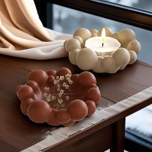 Puede incluir: Dos portavelas de cerámica con forma de nube. El portavelas de la izquierda es marrón y el de la derecha es beige. El portavelas beige tiene una vela blanca dentro.