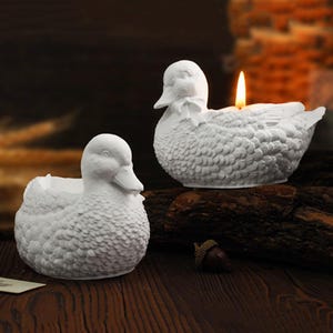Puede incluir: Dos portavelas de cerámica blanca con forma de pato. El pato de la derecha tiene una vela encendida dentro. Ambos patos tienen una superficie texturizada que se asemeja a plumas.