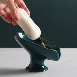 May include: A green ceramic soap dish with gold accents. The dish has a curved shape and a raised base. A bar of soap is being placed in the dish.