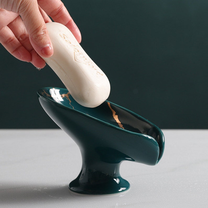 May include: A green ceramic soap dish with gold accents. The dish has a curved shape and a raised base. A bar of soap is being placed in the dish.