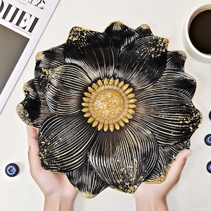 Peut inclure: Un plateau décoratif noir et doré en forme de fleur. Le plateau présente des motifs de pétales détaillés avec des accents dorés et un centre texturé. Le plateau est tenu par des mains sur un fond blanc. Une tasse de café et des myrtilles sont également visibles.