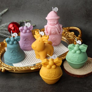 Puede incluir: Seis velas con temática navideña en diferentes formas y colores, incluyendo un muñeco de nieve, un reno y campanas. Las velas están decoradas con las palabras "Feliz Navidad".