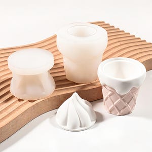 May include: A set of silicone moulds for crafting, including a cylindrical mould, a flat-topped circular mould, a swirled dome-shaped mould, and a small cup-shaped mould with a textured waffle pattern on the exterior and a smooth white rim.