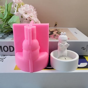 May include: Pink silicone mold for making a snowman candle and a white ceramic candle holder with a frog design. The frog is holding its hands up in a welcoming gesture.