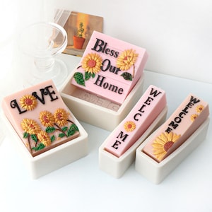May include: Four pink and white silicone molds for making soap bars. The molds are filled with pink soap bars decorated with sunflowers and the words "Love", "Welcome", "Bless Our Home", and "Welcome".