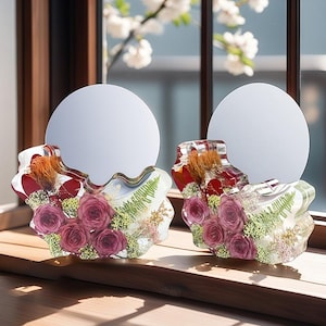 May include: Two clear resin flower arrangements with a round mirror in the center. Each arrangement features a cluster of pink roses and other dried flowers.
