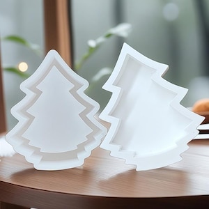 Puede incluir: Dos moldes blancos en forma de árbol de Navidad. Los moldes están hechos de un material translúcido y tienen un diseño en capas. Los moldes están sobre una mesa de madera. El fondo está borroso, con toques de vegetación.