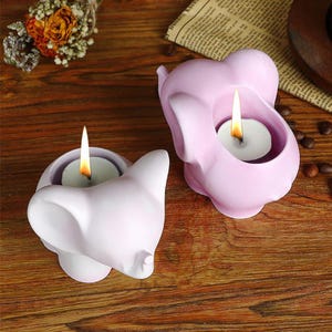 Może przedstawiać: Dwa różowe ceramiczne świeczniki w kształcie słonia z białą świeczką typu tealight w każdym. Świeczniki znajdują się na drewnianej powierzchni.