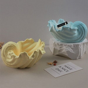 Może przedstawiać: Dwa ozdobne świeczniki w kształcie muszli. Świecznik po lewej stronie jest żółty, a świecznik po prawej stronie jest niebieski. Oba świeczniki mają fakturowany, falisty wzór. Niebieski świecznik ma małą czarną etykietę z napisem "HANDMADE".