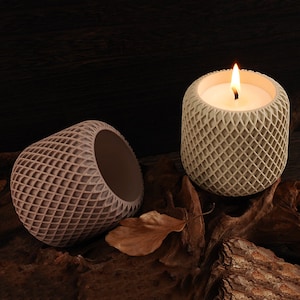 Op de afbeelding: Twee decoratieve kaarsen in een geometrisch patroon. De kaars rechts is aangestoken en heeft een beige kleur. De kaars links is niet aangestoken en heeft een bruine kleur.