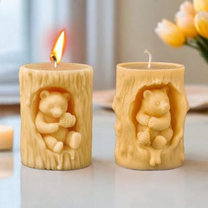 Puede incluir: Dos velas de pilar beige con un diseño de tronco de árbol y una figurita de oso dentro de cada una. Las velas están encendidas y tienen un brillo cálido.
