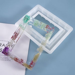 Puede incluir: Un molde de silicona blanco para hacer marcos de fotos rectangulares de resina. El molde tiene un borde elevado y una superficie lisa. El molde se muestra con un marco de resina terminado lleno de flores secas.