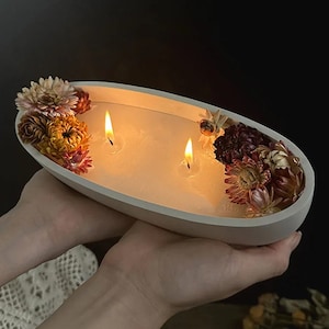 Könnte beinhalten: Eine weiße ovale Keramikschale mit drei brennenden Kerzen und getrockneten Blumen in Rosa-, Gelb- und Brauntönen.