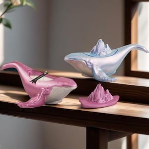 Puede incluir: Dos portavelas con forma de ballena con una montaña en la parte superior. La ballena de la izquierda es rosa y la ballena de la derecha es azul.