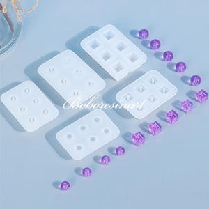 May include: Set of four silicone molds for making resin jewelry. The molds are white and feature different shapes, including circles and squares. There are also several purple resin beads scattered around the molds.