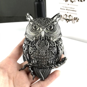 May include: A silver metal owl figurine with intricate details and a black finish. The owl is perched on a branch with its wings spread out.