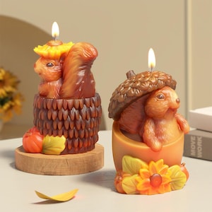 May include: Two autumn-themed candles shaped like squirrels. One squirrel is holding an acorn and the other is sitting on a bed of leaves. Both candles are decorated with fall colors and textures.
