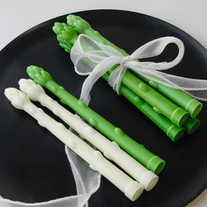 Op de afbeelding: Een set groene en witte kaarsen in de vorm van asperges. De kaarsen zijn met een wit lint aan elkaar gebonden en liggen op een zwart bord.