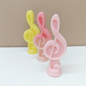 Puede incluir: Tres velas decorativas con forma de nota musical. Una es amarilla y dos son rosas.