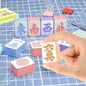 May include: A set of colourful mahjong tiles with Chinese characters on them. The tiles are arranged in a row on a blue grid surface. A hand is holding a tile with the character "萬" on it.