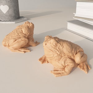 May include: Two beige ceramic frog figurines. The frogs are sitting on a white surface.