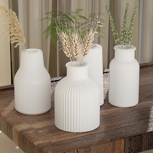 May include: Four white ceramic vases of different shapes and sizes, with a variety of dried flowers and grasses inside. The vases are arranged on a wooden table.
