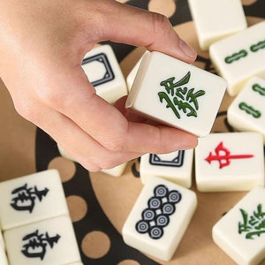 K&ouml;nnte beinhalten: Eine Hand h&auml;lt einen wei&szlig;en Mahjong-Stein mit gr&uuml;nen chinesischen Schriftzeichen. Andere Steine mit verschiedenen Symbolen und Zeichen sind verstreut. Die Steine liegen auf einer hellbraunen Oberfl&auml;che.