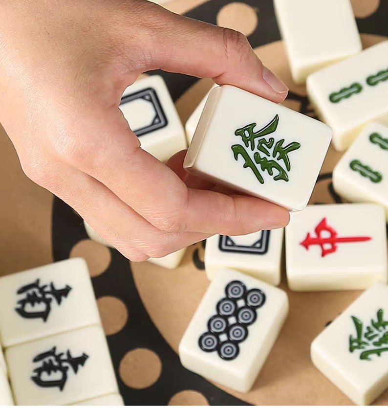 K&ouml;nnte beinhalten: Eine Hand h&auml;lt einen wei&szlig;en Mahjong-Stein mit gr&uuml;nen chinesischen Schriftzeichen. Andere Steine mit verschiedenen Symbolen und Zeichen sind verstreut. Die Steine liegen auf einer hellbraunen Oberfl&auml;che.