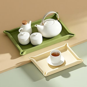 May include: A matte white ceramic teapot with a striped handle, a small pitcher with a cork stopper, and two lidded bowls arranged on a rectangular olive green tray, alongside a separate cream-colored tray holding a single white teacup on a saucer.