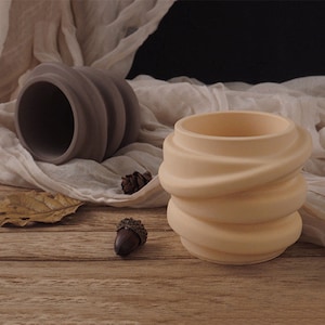 Puede incluir: Dos macetas de cerámica con un diseño en espiral. Una es beige, la otra gris. Las macetas están sobre una superficie de madera con una tela de color neutro en el fondo. También se ven una bellota y una hoja seca.