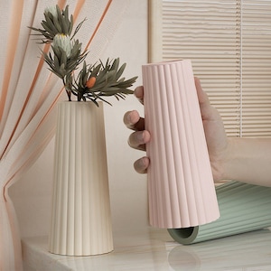 Könnte beinhalten: Zwei Keramikvasen mit geripptem Design. Eine Vase ist weiß und die andere rosa. Die Vasen stehen auf einer hellgrauen Oberfläche.