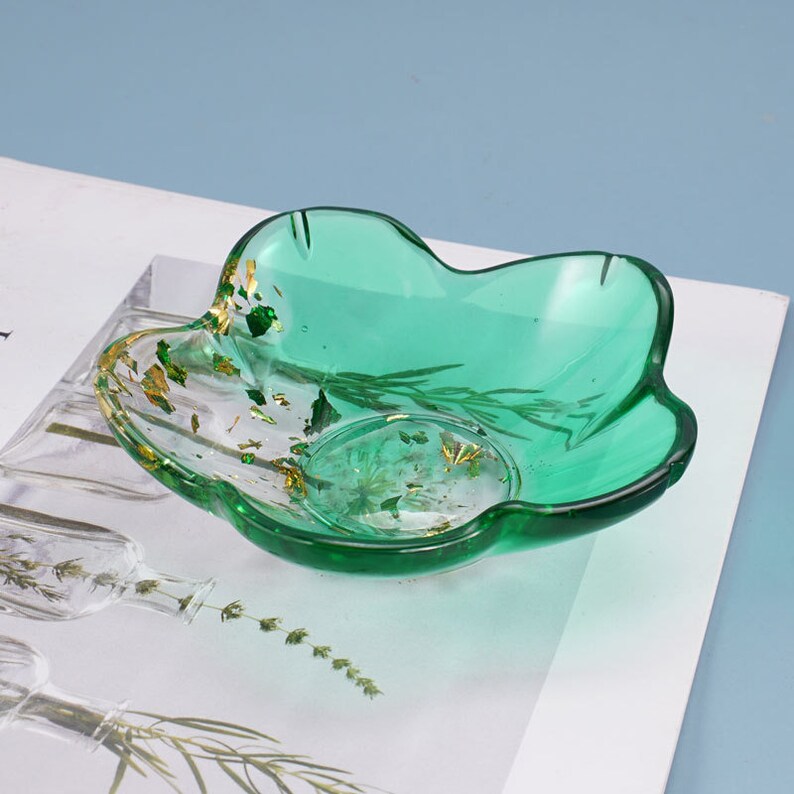 May include: A green resin trinket dish with a wavy edge and gold flecks. The dish has a clear center with dried flowers and leaves embedded in the resin.