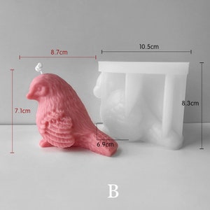 May include: A pink bird-shaped candle, 7.1 cm tall and 8.7 cm long, next to a white silicone mold. The mold is 8.3 cm tall and 10.5 cm wide. The candle has a wick and detailed feathering. The letter "B" is at the bottom.