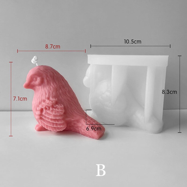 May include: A pink bird-shaped candle, 7.1 cm tall and 8.7 cm long, next to a white silicone mold. The mold is 8.3 cm tall and 10.5 cm wide. The candle has a wick and detailed feathering. The letter "B" is at the bottom.