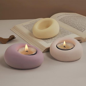 Pode incluir: Três castiçais modernos em cores pastel: lavanda, rosa claro e creme. Cada suporte tem um design circular com uma área rebaixada para uma vela de chá. Uma vela está acesa, emitindo um brilho quente. Os castiçais estão dispostos sobre uma mesa com um livro aberto.