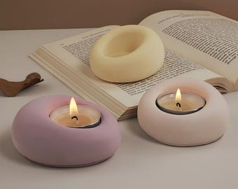 Puede incluir: Tres candelabros modernos en colores pastel: lavanda, rosa claro y crema. Cada soporte tiene un diseño circular con un área empotrada para una vela de té. Una vela está encendida, emitiendo un brillo cálido. Los candelabros están colocados sobre una mesa con un libro abierto.