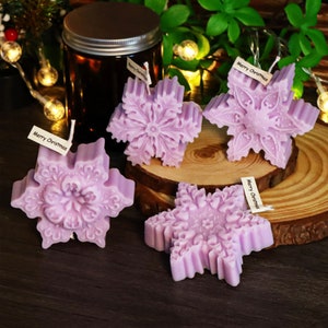 Può includere: Quattro candele viola a forma di fiocco di neve con il testo "Merry Christmas" su tre delle candele. Le candele sono disposte su una superficie di legno.