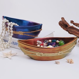 Op de afbeelding: Twee decoratieve keramische boten met gouden touwaccenten. De boot links is blauw en paars met witte parels erin. De boot rechts is oranje en goud met gekleurde kralen en een witte parelketting erin.