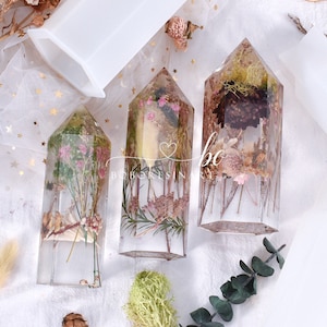 May include: Three clear resin crystal shapes filled with dried flowers and greenery. The crystals are in a hexagonal shape and are clear with a slight amber tint. The flowers are pink, white, and brown. The greenery is green and brown.