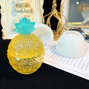 Puede incluir: Un molde de resina transparente en forma de piña con una parte superior turquesa. El molde está sobre una superficie blanca junto a un segundo molde de resina transparente más grande.