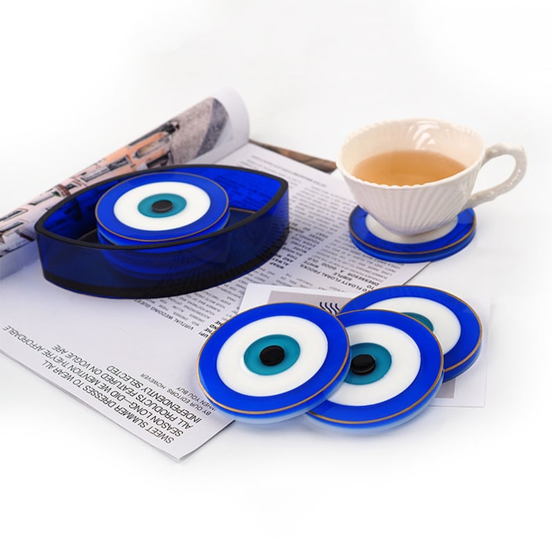 Op de afbeelding: Set van zes blauwe en witte onderzetters met gouden rand. De onderzetters zijn in een cirkel rond een witte theekop met een blauw boze oogontwerp gerangschikt. De onderzetters zijn gemaakt van glas en hebben een glanzende afwerking.
