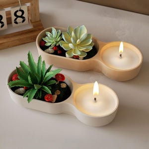 May include: Two white ceramic candle holders with a small succulent plant in one and a lit candle in the other. The candle holders are connected and have a rounded shape.