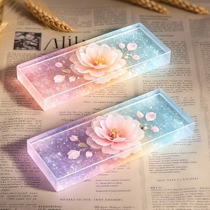 Puede incluir: Dos bandejas rectangulares de resina con un degradado de color que va del rosa suave al azul claro. Cada bandeja tiene incrustada una flor grande de color rosa pálido en el centro, rodeada de pétalos delicados y finas partículas de purpurina.