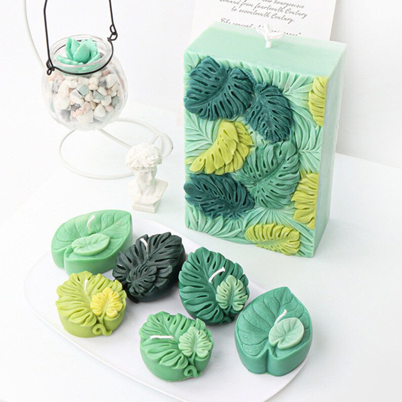 May include: A selection of green candles in different shapes, featuring a large rectangular candle with embossed monstera leaves and smaller leaf-shaped candles. The candles are in shades of green and yellow-green, creating a tropical theme.