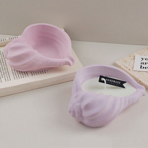Può includere: Due portacandele a forma di conchiglia di colore lavanda. Un portacandele contiene una candela bianca e un'etichetta nera su cui è scritto "HANDMADE" con un piccolo cartellino bianco che dice "specialmente per te".