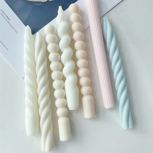 May include: Six decorative candles in various styles and colors. The candles are white, light blue, and light pink. Some candles have a twisted design, while others have a beaded design.