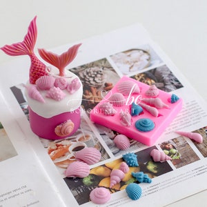 Könnte beinhalten: Rosa-weiße Meerjungfrauenkerze mit Muscheln und eine Silikonform mit Muschelformen. Die Form ist rosa und hat einen weißen Rand. Der Text "BOBORESINART" ist auf die Form gedruckt.